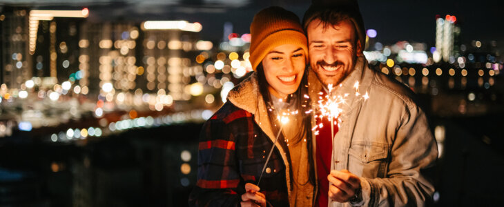 A couple holds sparklers on the rooftop of a building during New Year's Eve. City skyline behind them